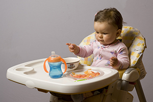 Bebé sentado en una silla alta con un vaso con popote y tratando de alcanzar bocados de alimento.