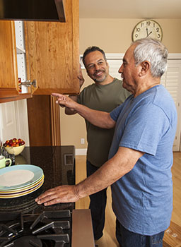 Older man standing in front of an open cupboard and pointing at it. A younger man stands next to him watching.